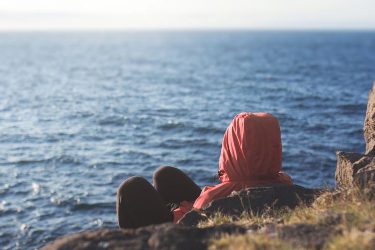 A Hooded Person Sitting on a Cliff Overseeing the Sea