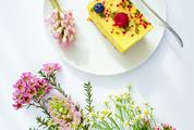Slice of Cheesecake Decorated with Fruits Surrounded by Flowers on White Table