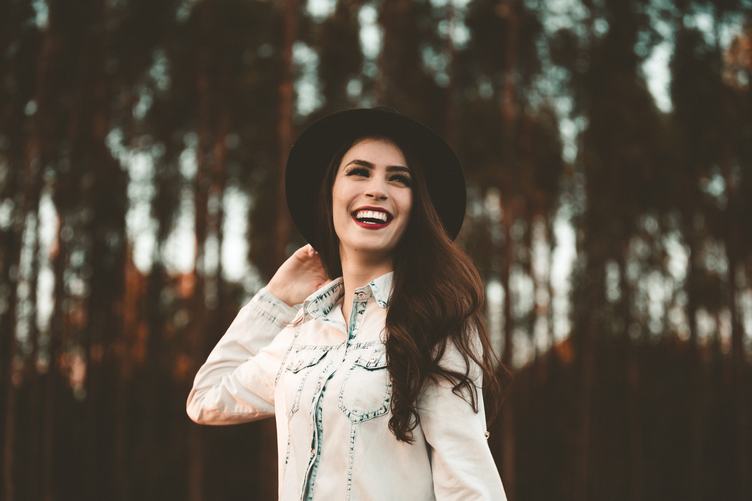 Stylish Young Woman Wearing Black hat Smiling against Blurred Trees