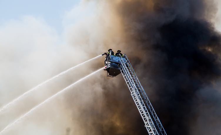 Two Firefmen on the Ladder Spray Water against Black Smoke